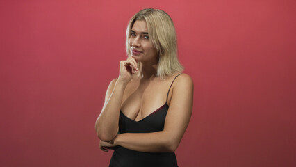 Fototapeta premium Young blonde woman touching chin with hand and crossed arms, wearing low-cut black tanktop against red studio backdrop; thoughtful contemplation.