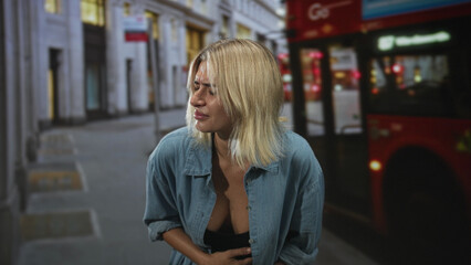 Fototapeta premium Young woman clutching stomach beside a red double decker bus on a busy street, grimacing with stomachache; sickness discomfort.