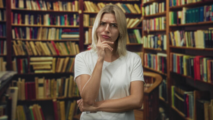 Young blonde woman with hand on chin and crossed arm wearing white tshirt beside tall crowded bookshelves in a library  pensive curiosity. © Krakenimages.com