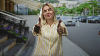 Fototapeta premium Woman smiling and giving thumbs up at hotel entrance by stairs and parked cars, doorman passing behind; optimism approval.