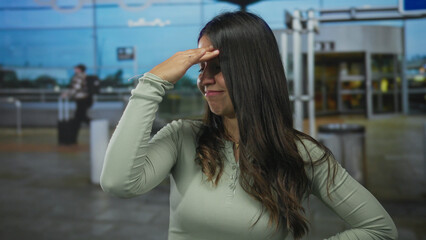 Fototapeta premium Young woman in airport terminal outdoors with a playful expression wearing a green shirt against a backdrop of busy travelers and airport signage.