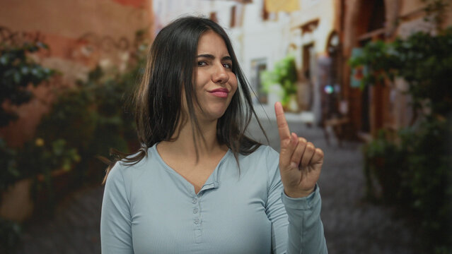 Young woman makes finger pointing gesture to herself on a narrow street in soft daylight; rejection.