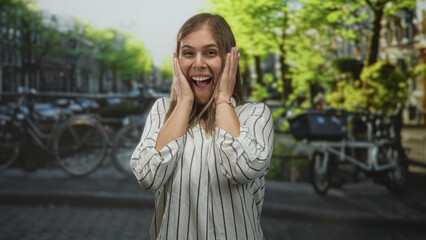Fototapeta premium Woman hands to cheeks smiling on sunlit street in amsterdam canal area wearing striped blouse and watch; surprise travel moment.