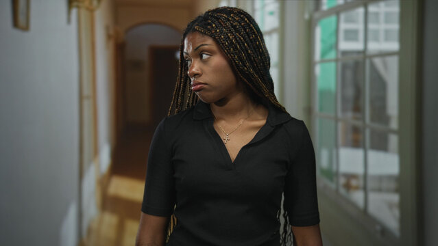 African american woman face looking forward in hotel corridor, standing with neutral expression and braided hair; quiet disappointment.