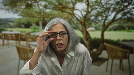 Woman grey haired adjusting eyeglasses while squinting and leaning forward on a restaurant terrace outdoors; curiosity reflection.