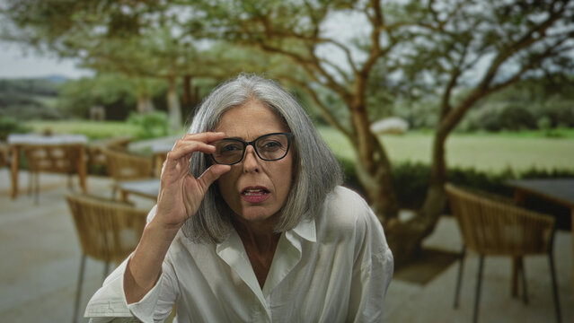 Woman grey haired adjusting eyeglasses while squinting and leaning forward on a restaurant terrace outdoors; curiosity reflection.