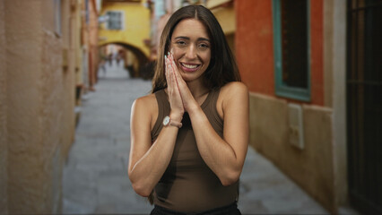 Naklejka premium Young hispanic woman clapping hands and smiling on a sunlit narrow street with colorful buildings and archway, wearing brown tank top; joy.
