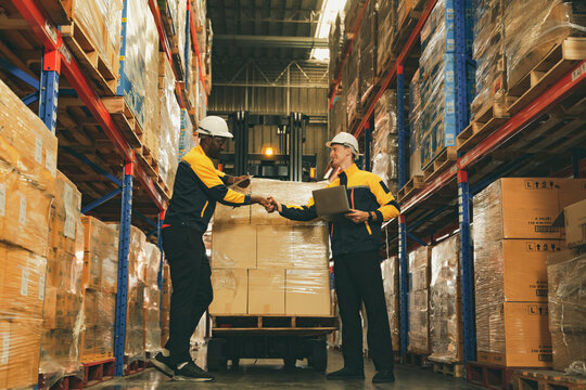 Two male workers shake hands as colleagues warehouse employees responsible for inspecting goods and operating forklift demonstrating teamwork and positive camaraderie at work among foreign workers.