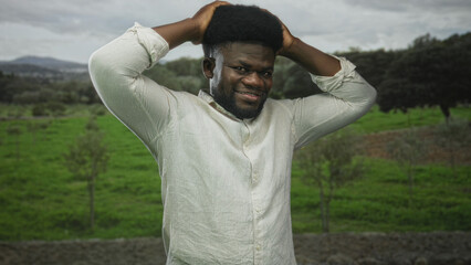 Man in white shirt smiling with hands on head, afro hair visible, standing in a forest field of green grass under a cloudy sky; serenity relaxation.