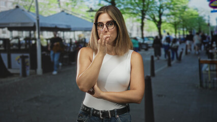 Fototapeta premium Woman wearing glasses and a white sleeveless top touches her lips with fingers in a finger to lips gesture, arms crossed while standing on a busy street; hush contemplation.