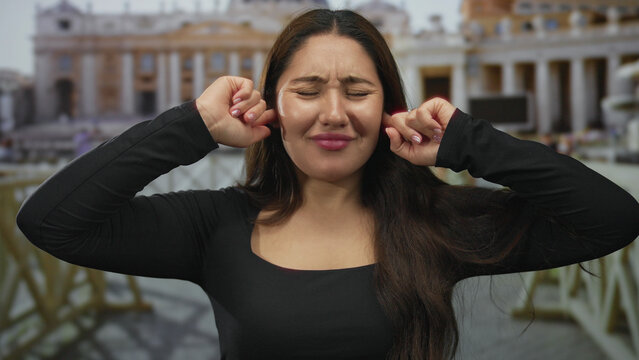 Young hispanic woman covers ears with both hands and closes eyes in vatican square amid st peter building facade; annoyance.