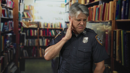 Police man presses hand to ear beside crowded bookshelves in library building, radio on shoulder and badge visible  pain. © Krakenimages.com