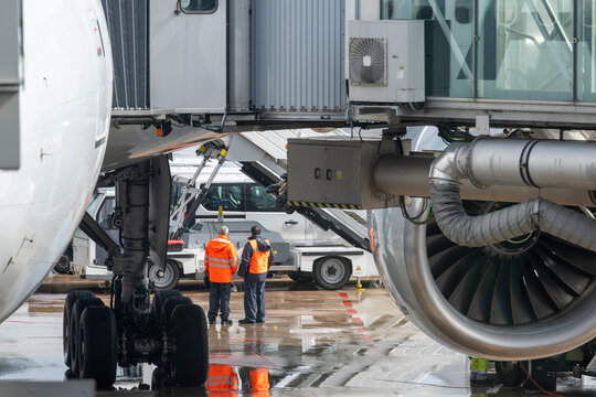 Airport workers standing beside aircraft engine and landing gear while discussing ground service operations near terminal jet bridge.