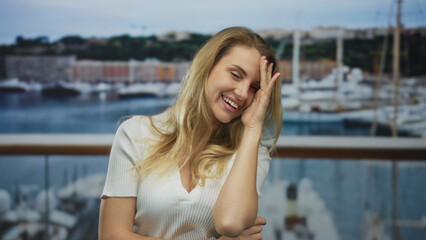 Obraz premium Woman smiling with hand on forehead at port with boats and sea in the background, conveying a joyful outdoor moment.