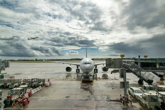 Front view of passenger airplane connected to pushback tractor on airport apron while another aircraft takes off under dramatic cloudy sky.