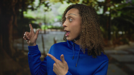 Woman wearing bright blue hoodie points finger to side near tree trunk in green outdoor park  enthusiasm. © Krakenimages.com