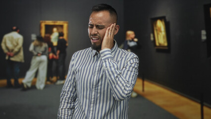 Obraz premium Man holding hand to cheek and wincing in an art gallery building with framed paintings and visitors nearby; toothache discomfort.
