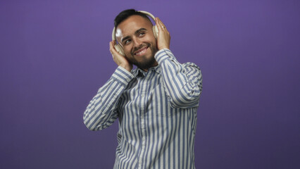 Man smiling with closed eyes holding headphones over ears and touching earcups in purple studio while wearing a striped shirt; joy relaxation.
