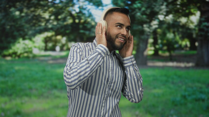 Man with headphones and striped shirt giving thumbs up while smiling in a forest park with trees...