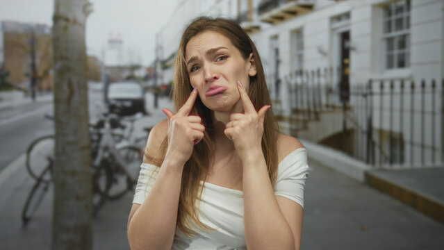 Young caucasian woman mimes crying tears with fingers on urban street sidewalk near bicycles and row houses; sarcasm.