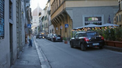 Obraz premium Narrow historic street with parked cars and lit cafe terrace, softly defocused bokeh street; background copyspace backplate calm.