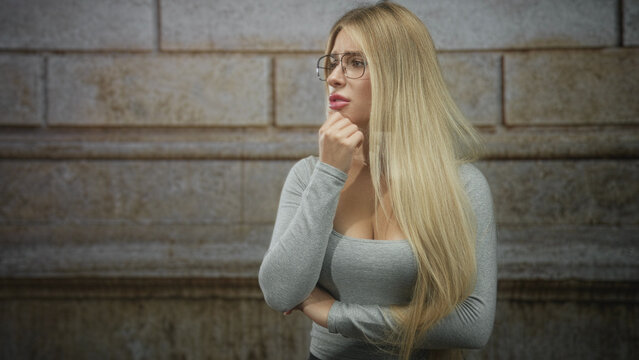 Woman wearing glasses with hand on chin and folded arm, long blonde hair, grey fitted top showing cleavage, standing before rusticated stone building wall; pensive.