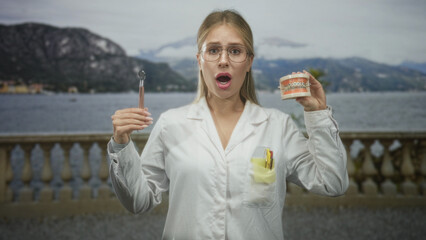 Woman dentist in white coat holding dental mirror and braces model, mouth open on building balcony;...