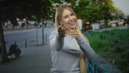Young blonde woman in long sleeve top pointing both fingers like finger guns at camera on street beside graffiti wall and parked scooter; playful confidence.