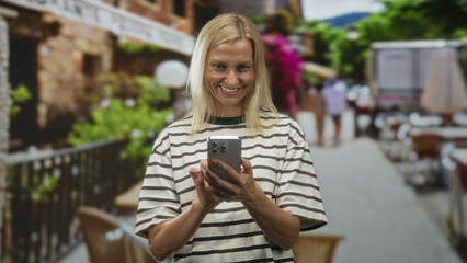 Woman blonde smiling, tapping smartphone with thumbs, wearing striped shirt in a busy street cafe;...