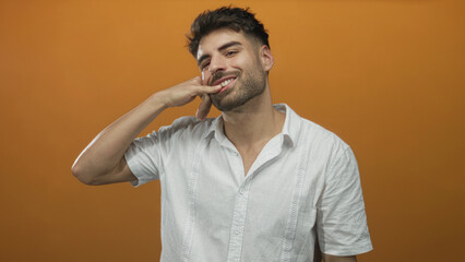 Obraz premium Man makes call gesture in orange studio background wearing white shirt, looking at hand; invitation.
