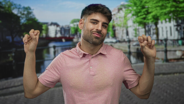 Young hispanic man in pink polo shirt showing playful smirk with hands making quote gesture on a street; sarcasm.