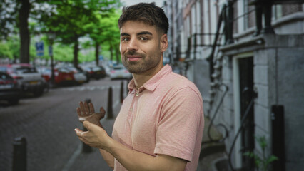Young hispanic man gestures with open hands on bustling city street beneath green trees wearing pink polo; curiosity.