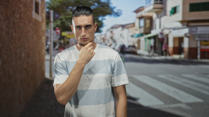 Fototapeta premium Man with hand on chin at pedestrian crosswalk on sunlit street near storefronts and buildings; contemplation thoughtful.