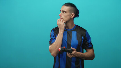 Man in blue soccer jersey holding a gamepad with fingers to mouth and looking sideways in studio  contemplation focus. © Krakenimages.com