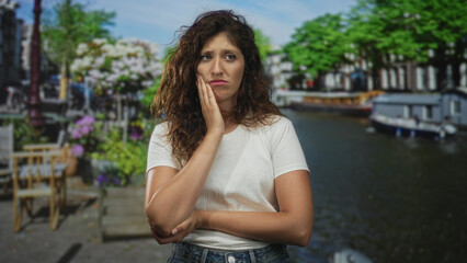 Fototapeta premium Woman touching chin with hand, freckled face and folded arm, wearing white tee, standing on canal street by houseboats in amsterdam; thoughtful reflection.
