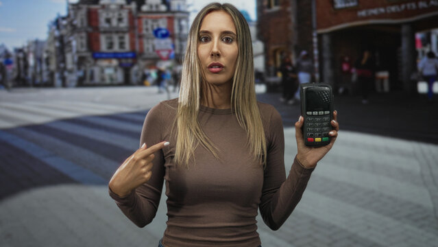 Woman points finger to card reader while holding payment terminal on a busy city street near shops; payment confusion.