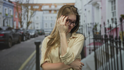 Obraz premium Young blonde woman tucks hair behind ear on urban street in front of pastel buildings with slight smile; shyness.