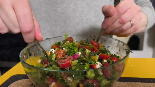 A young man preparing a fresh vegetable salad at home, gently mixing colorful ingredients and leafy greens with two spoons in a large transparent glass bowl. Healthy lifestyle, home cooking, and fresh