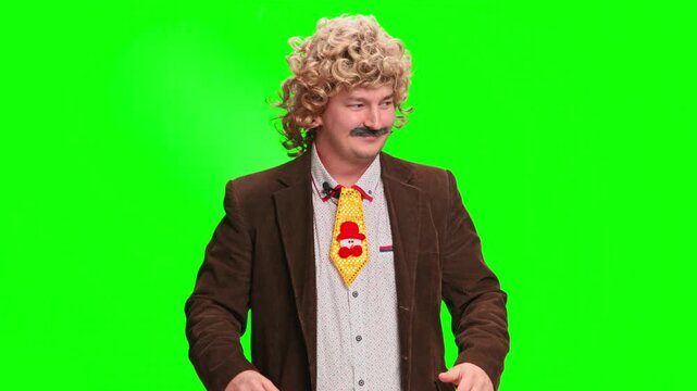 A grown man in a curly blond wig and false mustache, wearing a brown jacket and an unusual tie, stands in front of a bright green screen, fidgeting and preparing to give a speech.
