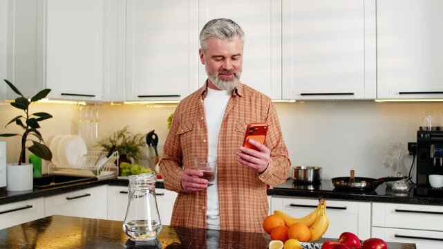 Middle-aged man smiles reading smartphone while drinking water in home kitchen during morning diet focus. Mature guy tracks hydration and healthy eating goals using mobile app indoors with calm mood.