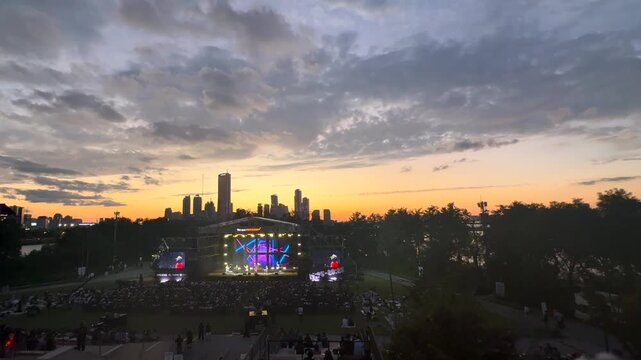 Large outdoor music festival at sunset with crowd watching illuminated concert stage in city park, downtown skyline in background during evening live entertainment event.