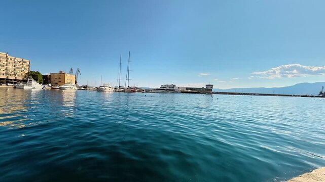 A panoramic daytime view of the Pula waterfront from a concrete pier. The deep blue Adriatic Sea fills the foreground, with various yachts, sailboats, and harbor buildings lining the coast against a