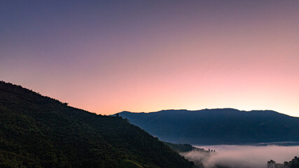 Fototapeta premium Scenic drone view of a misty mountain valley at sunrise in Sapan Village, Nan Thailand. A small village glows beneath drifting fog and soft golden light, showcasing a peaceful countryside travel