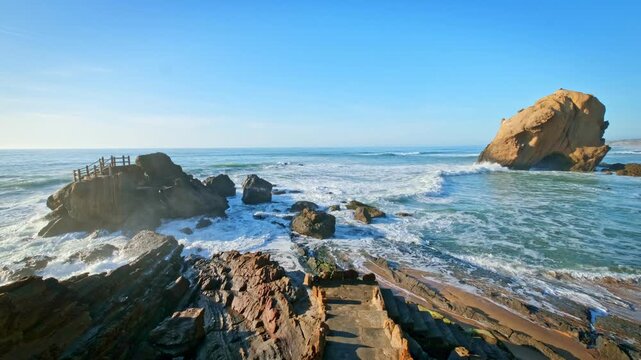 Penedo do Guincho at Praia da Santa Cruz in Portugal displays a rock arch with waves breaking on the coast. Visitors walk along the sandy beach enjoying the view of the ocean.