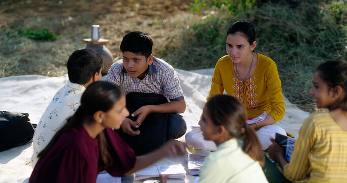 Indian villager girl boy sitting on mat do study read book outdoor rural area ground place. Happy group child hold notes talk gossip enjoy desi life new day outside open class field