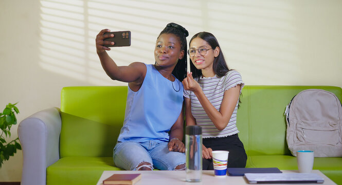 Happy young adult gen z woman sitting on sofa couch use mobile look cam take selfie indoor lounge room place teen age girl hold phone record casual video spend break time do funny pose enjoy new day