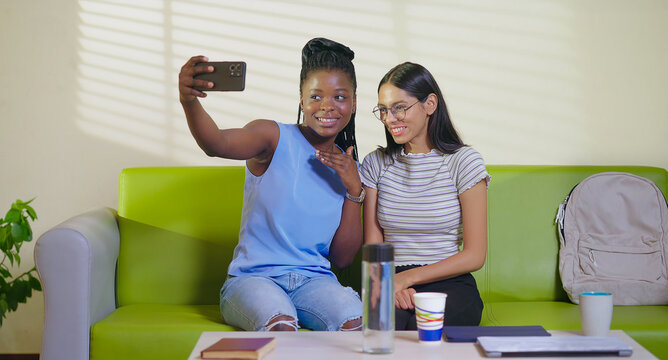 Happy young adult gen z woman sitting on sofa couch use mobile look cam take selfie indoor lounge room place teen age girl hold phone record casual video spend break time do funny pose enjoy new day