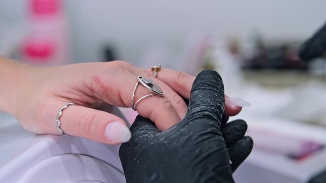 Salon manicure detail showing black gloved hands refining the edge of a long fingernail with an emery board while supporting the hand.