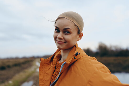 Portrait of a smiling woman outdoors wearing an orange jacket and a wig cap. The background shows a cloudy sky and natural landscape. Concept of woman lifestyle and confidence.
