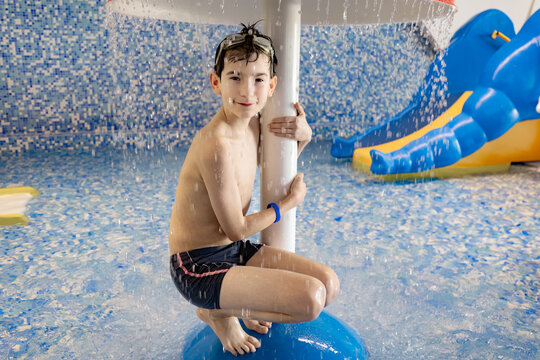 Young boy in swim shorts enjoys water play under a fountain at an indoor water park, colorful slides and tiled walls visible in the background